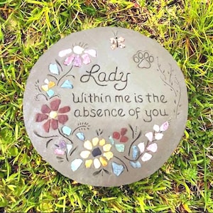 May include: A round, gray stone garden stepping stone with the words "Lady Within me is the absence of you" in black script. The stone is decorated with colorful mosaic flowers, a butterfly, and a paw print.