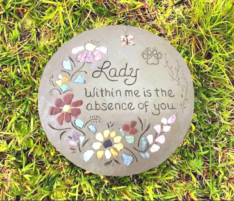 May include: A round, gray stone garden stepping stone with the words "Lady Within me is the absence of you" in black script. The stone is decorated with colorful mosaic flowers, a butterfly, and a paw print.