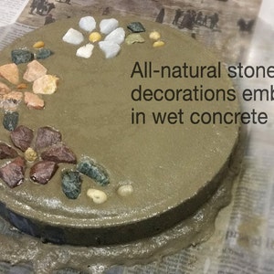May include: A round, gray concrete stepping stone decorated with colorful natural stones. The text "All-natural stone decorations embedded in wet concrete" is visible. The stones are arranged in floral patterns.