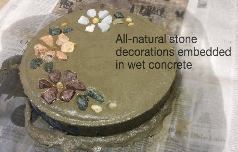 May include: A round, gray concrete stepping stone decorated with colorful natural stones. The text "All-natural stone decorations embedded in wet concrete" is visible. The stones are arranged in floral patterns.