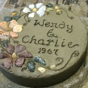 May include: A round, gray concrete stepping stone with the names "Wendy & Charlie" and the year "1967" inscribed. The stone is decorated with colorful stones arranged in floral patterns.