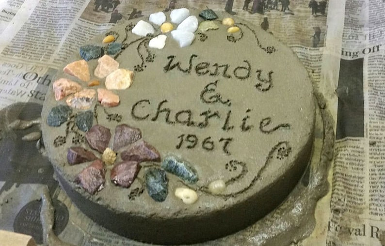 May include: A round, gray concrete stepping stone with the names "Wendy & Charlie" and the year "1967" inscribed. The stone is decorated with colorful stones arranged in floral patterns.