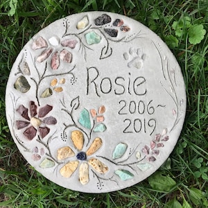 May include: A round, gray concrete garden stone with floral and paw print designs. The name "Rosie" and the years 2006-2019 are inscribed. The flowers are made of colorful stones, and the stone is set in green grass.
