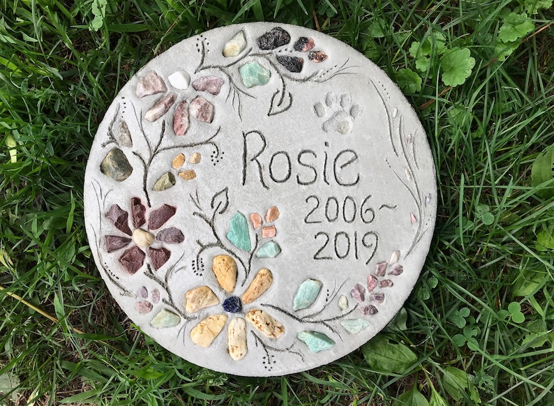 May include: A round, gray concrete garden stone with floral and paw print designs. The name "Rosie" and the years 2006-2019 are inscribed. The flowers are made of colorful stones, and the stone is set in green grass.