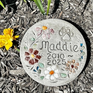 May include: A round, gray stone garden marker with the name "Maddie" and the years 2010-2025. The stone is decorated with colorful inlaid stones in a floral design and a paw print. A yellow flower is visible in the background.