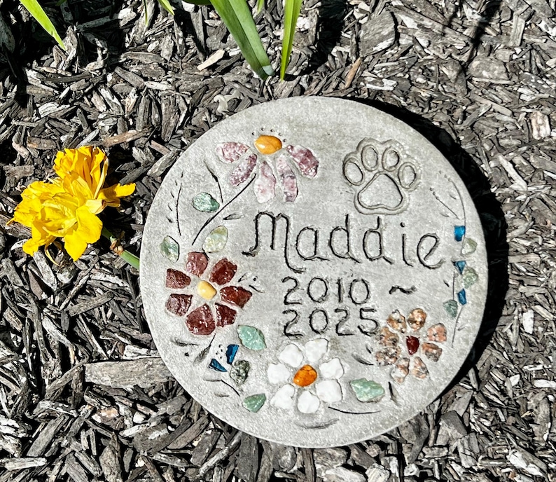 May include: A round, gray stone garden marker with the name "Maddie" and the years 2010-2025. The stone is decorated with colorful inlaid stones in a floral design and a paw print. A yellow flower is visible in the background.
