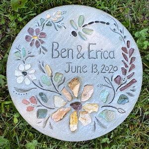 May include: A gray stone garden stepping stone with a mosaic design of flowers, leaves, and a dragonfly. The stone is personalized with the text "Ben & Erica June 13, 2020".