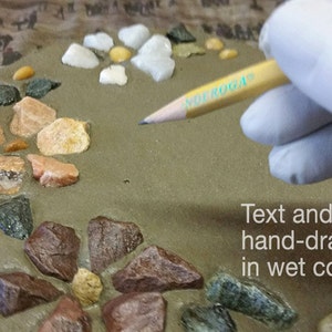 May include: A close-up of a concrete surface decorated with colorful stones. A hand in a white glove holds a pencil, drawing text and patterns on the wet concrete. The text reads: "Text and pattern hand-drawn in wet concrete."