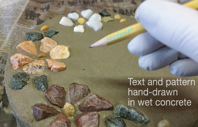 May include: A close-up of a concrete surface decorated with colorful stones. A hand in a white glove holds a pencil, drawing text and patterns on the wet concrete. The text reads: "Text and pattern hand-drawn in wet concrete."