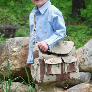 Puede incluir: Un niño pequeño con una camisa azul claro abotonada y pantalones caqui lleva una bolsa de mensajero de camuflaje con una correa de cuero marrón.