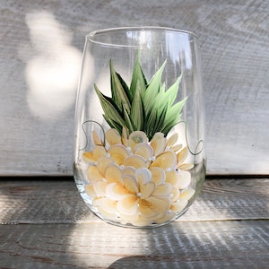 May include: A clear glass wine glass with a hand-painted pineapple design. The pineapple is yellow and white with green leaves.