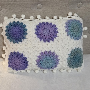 May include: A rectangular, white crochet pillow featuring six colorful floral designs in shades of purple and teal. The pillow is trimmed with white pom-poms along the edges, creating a decorative and textured appearance.