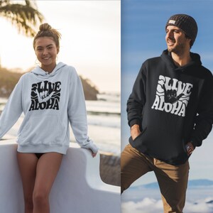 May include: Two people wearing black and white hoodies with a graphic of a hand making the shaka sign and the text "Live Aloha" in a retro font.