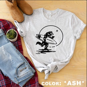 Puede incluir: Una camiseta gris con un gráfico negro de un oso surfeando en una ola con una luna llena y una estrella en el fondo. El texto "COLOR: *ASH*" está impreso debajo de la imagen.