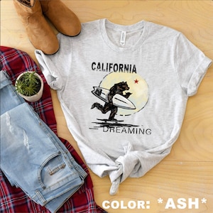 May include: A gray t-shirt with a graphic of a bear surfing with the text "CALIFORNIA DREAMING" printed on it. The text "COLOR: *ASH*" is printed at the bottom of the image.