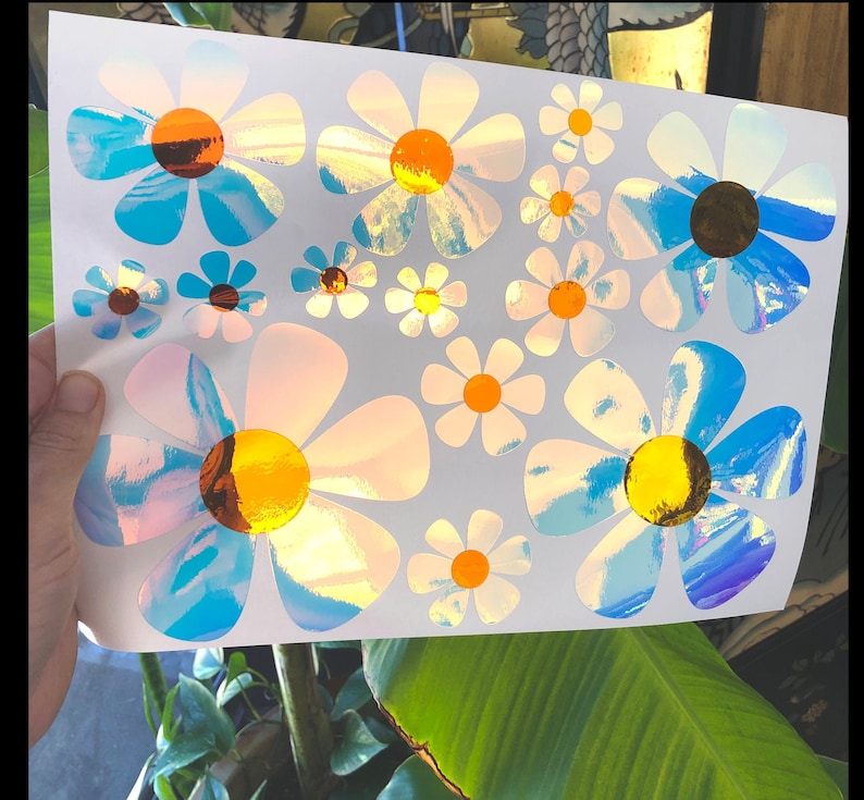 May include: A sheet of iridescent blue and white daisy stickers. The daisies have yellow centers and are arranged in a random pattern.