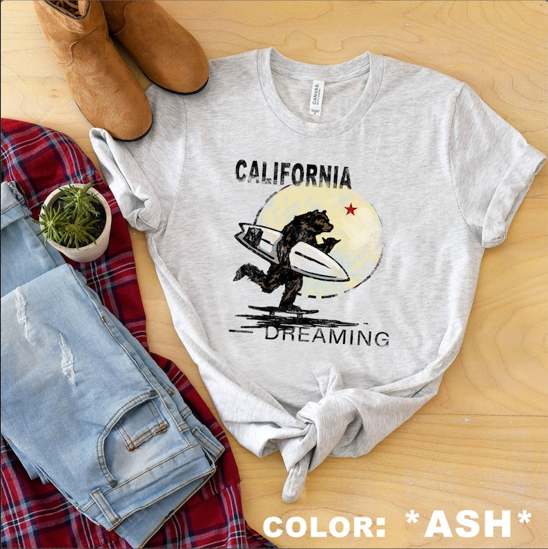 May include: A gray t-shirt with a graphic of a bear surfing with the text "CALIFORNIA DREAMING" printed on it. The text "COLOR: *ASH*" is printed at the bottom of the image.