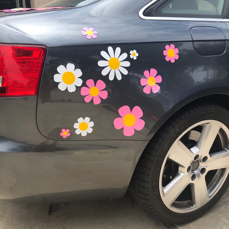 May include: A gray car with a variety of pink and white daisy stickers on the back bumper. The daisies have yellow centers.