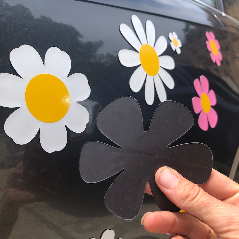 May include: A black flower-shaped magnet with five petals. The magnet is being held by a hand in front of a car with white daisy stickers.