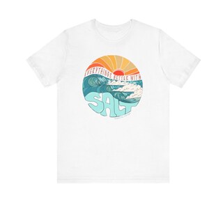 Puede incluir: Camiseta blanca con un gr&aacute;fico de una puesta de sol sobre olas oce&aacute;nicas. El gr&aacute;fico est&aacute; en un c&iacute;rculo con el texto "Everything's Better With Salt" alrededor de la parte superior y la palabra "Salt" en letras grandes y estilizadas en el centro.