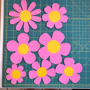 May include: Six pink paper flowers with yellow centers. The flowers are cut out and arranged on a green cutting mat.