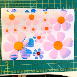 May include: A sheet of white paper with multiple iridescent pink and blue daisy stickers. The daisies have orange centers and are various sizes.