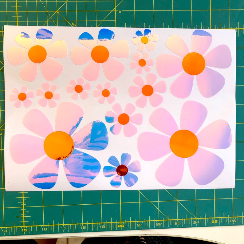 May include: A sheet of white paper with multiple iridescent pink and blue daisy stickers. The daisies have orange centers and are various sizes.