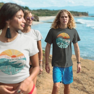 Puede incluir: Tres personas con camisetas con un gr&aacute;fico retro de un sol poni&eacute;ndose sobre las olas y la palabra "SALT" en un c&iacute;rculo. El gr&aacute;fico est&aacute; en tonos de azul, naranja y amarillo. Las personas caminan por una playa.