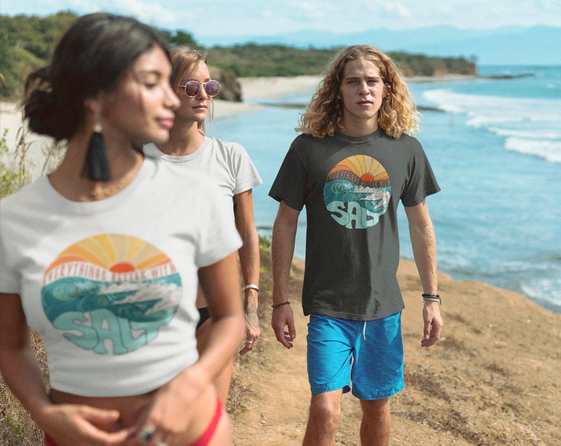 Puede incluir: Tres personas con camisetas con un gr&aacute;fico retro de un sol poni&eacute;ndose sobre las olas y la palabra "SALT" en un c&iacute;rculo. El gr&aacute;fico est&aacute; en tonos de azul, naranja y amarillo. Las personas caminan por una playa.