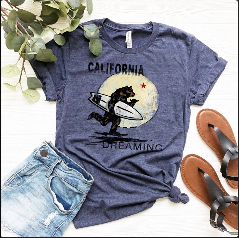 May include: A navy blue t-shirt with a graphic of a bear surfing on a wave with a yellow moon and the text "CALIFORNIA DREAMING" printed on the front.