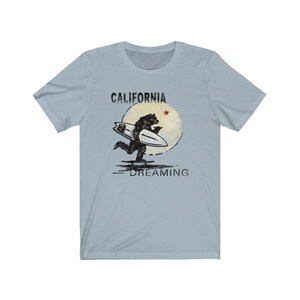 May include: A light blue t-shirt with a graphic of a bear surfing on a wave with a red star and the text "CALIFORNIA DREAMING".
