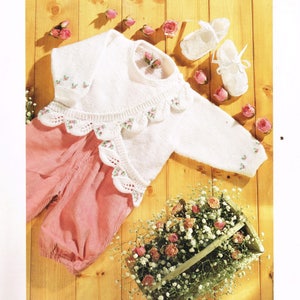 May include: A white knitted baby cardigan with a scalloped edge and floral embroidery. The cardigan is laid out on a wooden surface with pink baby pants and a basket of flowers.