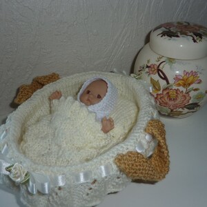 Puede incluir: Un moisés de bebé blanco de crochet con una muñeca dentro. El moisés tiene asas de crochet marrón y detalles de cinta de satén blanca. La muñeca lleva un gorro de crochet blanco y un atuendo de punto blanco.