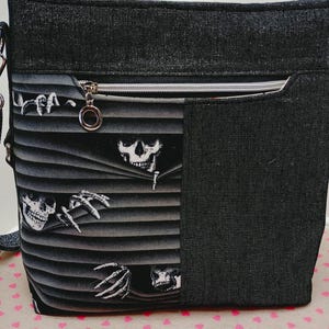 May include: Black tote bag with a zipper closure and a shoulder strap. The bag features a unique design with skulls and skeletal hands peeking through gray blinds. The bag is made of a textured black fabric.