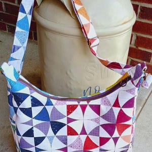 May include: A shoulder bag featuring a white fabric base with a repeating geometric pattern of multicolored curved shapes in shades of blue, purple, red, and orange. The bag includes an adjustable strap and a top zipper closure with metal hardware.