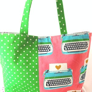 May include: Green and pink tote bag with a polka dot pattern on one side and a typewriter print on the other side. The typewriter print features a blue typewriter with a gold heart on a white piece of paper.