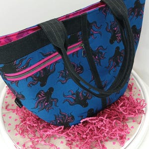 May include: A blue tote bag with a black octopus print and pink accents. The bag has two black straps and a zippered pocket on the front. The bag is sitting on a pink and white surface.