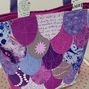 May include: A purple and gray tote bag with a patchwork design of overlapping scales. The bag has two purple straps and is made of fabric with a white background and black text.