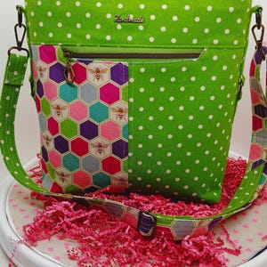 May include: A handmade green polka dot crossbody bag with a colorful bee and honeycomb pattern on the front pocket. The bag has a zipper closure, adjustable strap, and a small "Handmade" tag.
