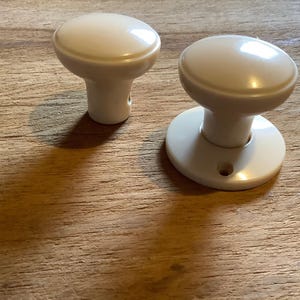 May include: Two white ceramic door knobs. One knob is round and has a flat base. The other knob is round and has a short, cylindrical base.