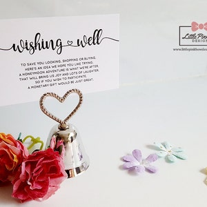 May include: A silver bell with a heart-shaped holder, used for wedding wishing well cards. The card reads "wishing well" and includes a message about a honeymoon adventure. The card also includes the website address for Little Pink Bow Designs.