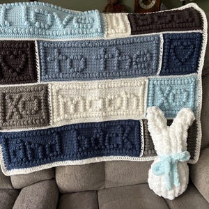 May include: A handmade crochet blanket in shades of blue, gray, and white, with the words "I LOVE YOU TO THE MOON AND BACK" stitched across it. A white plush bunny with a blue ribbon sits next to the blanket on a gray couch.