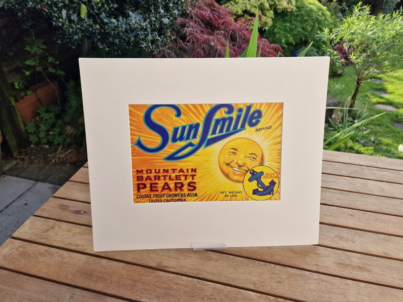 May include: Vintage Sun Smile brand label for Mountain Bartlett Pears. The label features a smiling sun face with a blue anchor logo and text that reads "Colfax Fruit Growers Assn. Colfax, California".