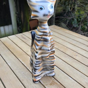 May include: A ceramic figurine of a cat with black, white and orange stripes. The cat has blue eyes and a pink nose. The figurine is standing on a wooden surface.