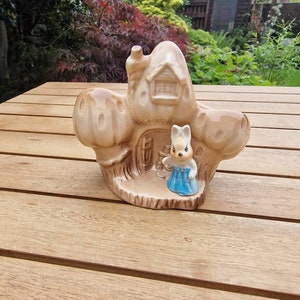May include: A ceramic figurine of a rabbit in a blue dress standing in front of a brown and white cottage with a chimney. The cottage has a small window and a door. The figurine is on a wooden table.