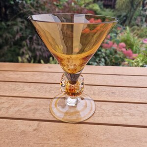 Puede incluir: Una copa de vidrio ámbar transparente con un cuenco ancho y un tallo decorativo. El tallo está hecho de vidrio transparente y tiene una serie de pequeñas cuentas redondas de color ámbar.