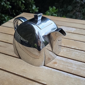 May include: A polished silver-tone metal teapot with a rounded, dome-shaped body, a curved handle, a short spout, and a square-shaped lid knob. The reflective surface shows a distorted view of the surroundings on its smooth, curved exterior.