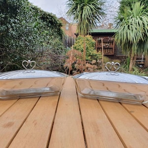 Könnte beinhalten: Zwei transparente Glas-Backformen mit Deckeln. Die Deckel haben silberne Griffe mit herzförmigem Design.