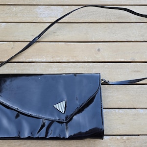 May include: A black patent leather clutch with a long, thin strap. The flap closure features a silver and white triangle emblem. The bag is rectangular with a curved flap and is set against a wooden background.