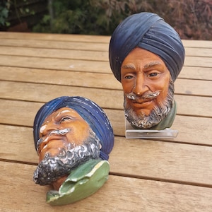 May include: Two decorative wall plaques featuring the heads of men wearing blue turbans. The plaques are made of ceramic and have a realistic painted finish.